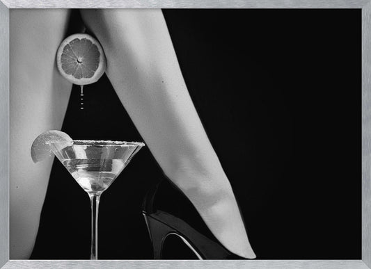 A framed, black and white photograph showing a woman's legs and a martini glass against a black background. A sliced citrus fruit is held between her thighs, with juice dripping towards the sugar-rimmed cocktail glass below. One foot is visible, wearing a black stiletto high heel. Artwork