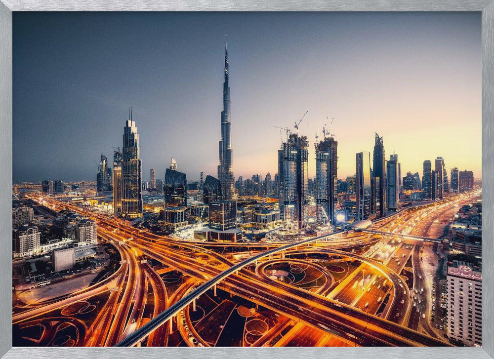 A wide-angle, elevated photograph of the Dubai skyline at dusk, with the iconic Burj Khalifa towering in the center. In the foreground, a complex network of highways glows with the orange light trails of moving traffic, while the city's numerous skyscrapers are illuminated against a twilight sky. Print