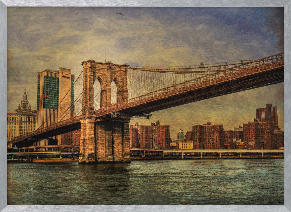 A framed, vintage-style photograph of the Brooklyn Bridge from a low angle, showing its stone pylon and suspension cables against a textured, cloudy sky. The Manhattan skyline and the East River are visible in the background and foreground. Wall Art