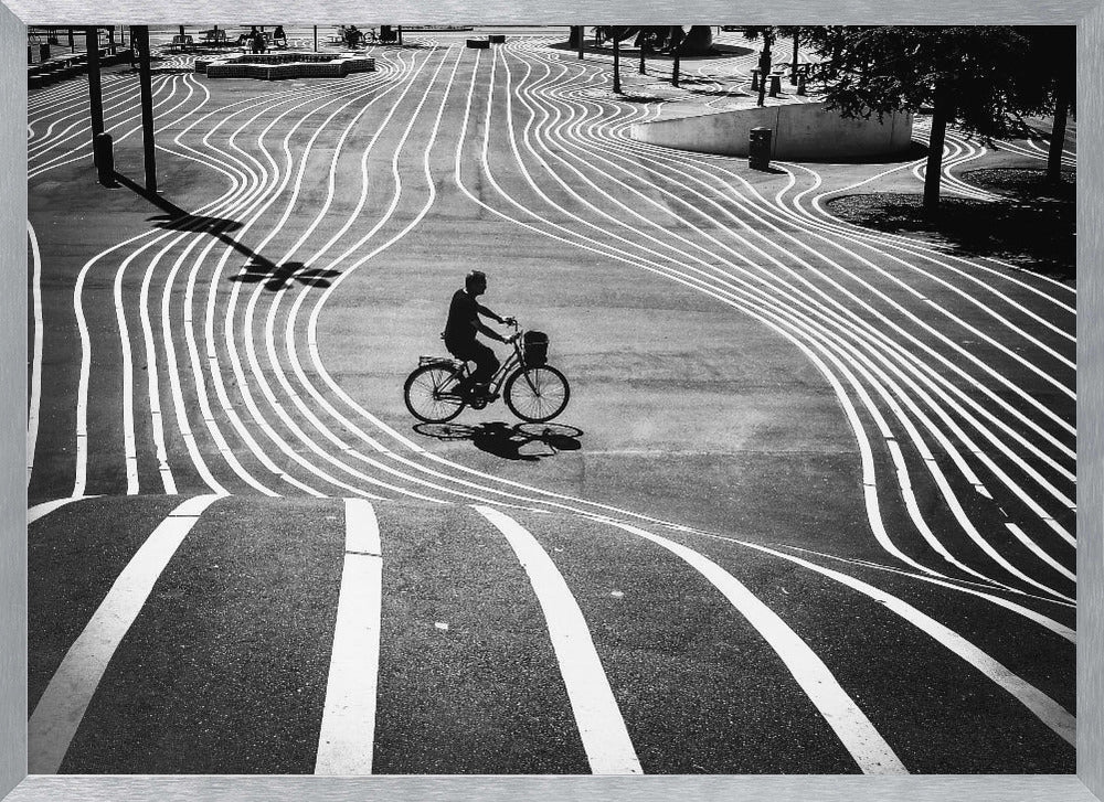A high-contrast, black and white overhead photograph of a person riding a bicycle across a wide, paved plaza. The ground is covered in a mesmerizing pattern of wavy, parallel white lines, and the cyclist's long shadow is cast by the bright sun. Print