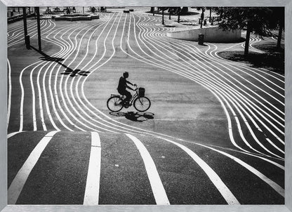A high-contrast, black and white overhead photograph of a person riding a bicycle across a wide, paved plaza. The ground is covered in a mesmerizing pattern of wavy, parallel white lines, and the cyclist's long shadow is cast by the bright sun. Print