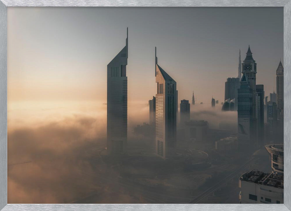 A framed aerial photograph of a futuristic city skyline, likely Dubai, enveloped in a thick layer of fog at sunrise. Tall, modern skyscrapers, including the iconic Emirates Towers, pierce through the clouds, with the warm light of the rising sun creating a hazy, golden glow on the left. Poster