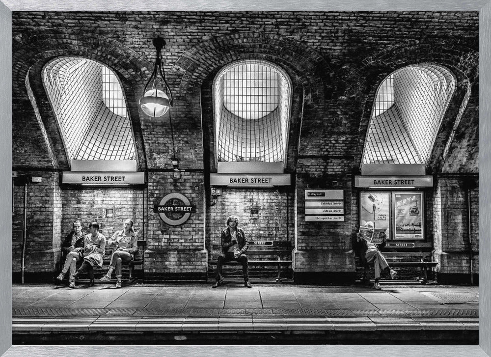 A framed, dramatic black and white photograph of the Baker Street underground station platform in London. Passengers wait on benches beneath iconic brick arches and large grated windows, with signs for 'Baker Street' and the London Underground roundel clearly visible. Print