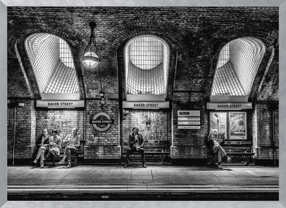 A framed, dramatic black and white photograph of the Baker Street underground station platform in London. Passengers wait on benches beneath iconic brick arches and large grated windows, with signs for 'Baker Street' and the London Underground roundel clearly visible. Print