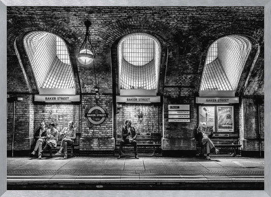 A framed, dramatic black and white photograph of the Baker Street underground station platform in London. Passengers wait on benches beneath iconic brick arches and large grated windows, with signs for 'Baker Street' and the London Underground roundel clearly visible. Print