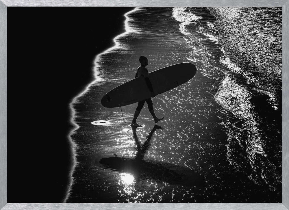 A dramatic, high-contrast, black and white aerial view of a silhouetted surfer carrying their surfboard along the shoreline. The sun glints off the wet sand and the dark water, creating a sparkling effect and a clear reflection of the surfer. Poster