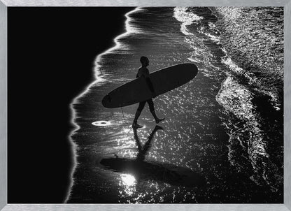 A dramatic, high-contrast, black and white aerial view of a silhouetted surfer carrying their surfboard along the shoreline. The sun glints off the wet sand and the dark water, creating a sparkling effect and a clear reflection of the surfer. Poster