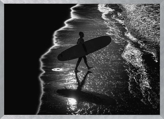 A dramatic, high-contrast, black and white aerial view of a silhouetted surfer carrying their surfboard along the shoreline. The sun glints off the wet sand and the dark water, creating a sparkling effect and a clear reflection of the surfer. Poster