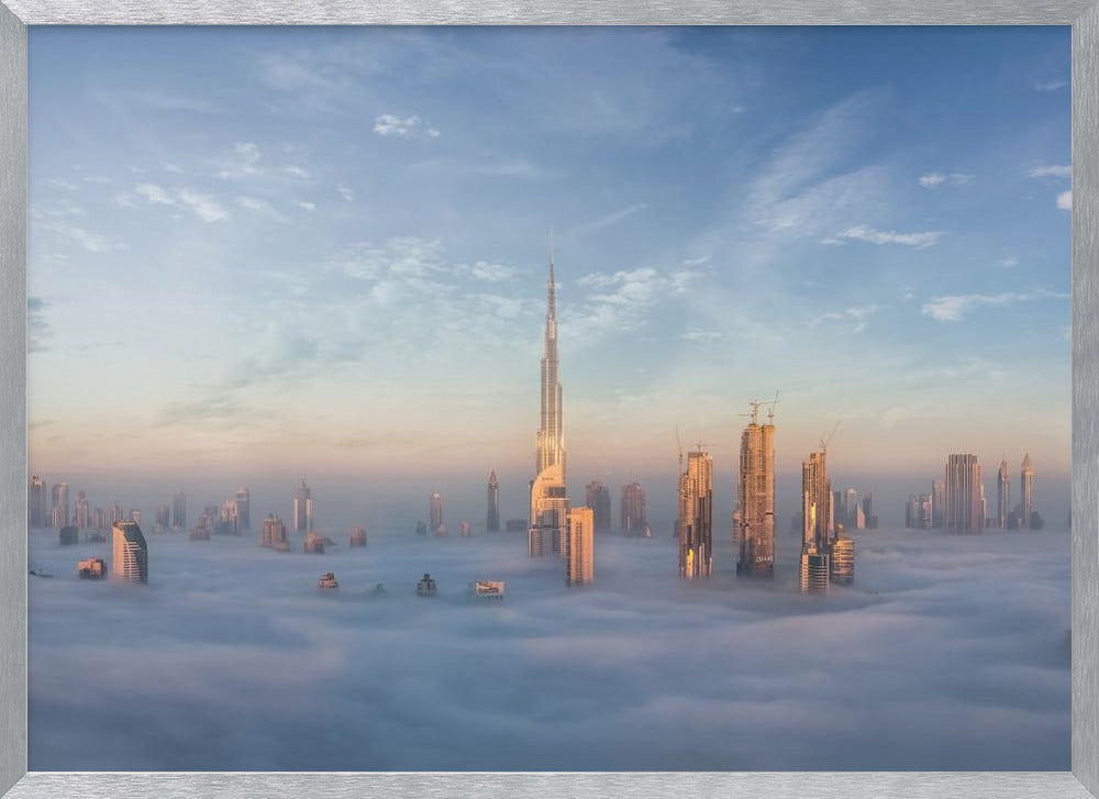A framed photograph of the Dubai skyline, with the Burj Khalifa and other skyscrapers piercing through a thick blanket of low-lying clouds or fog. The scene is bathed in the warm, golden light of sunrise or sunset against a pale blue sky. Poster
