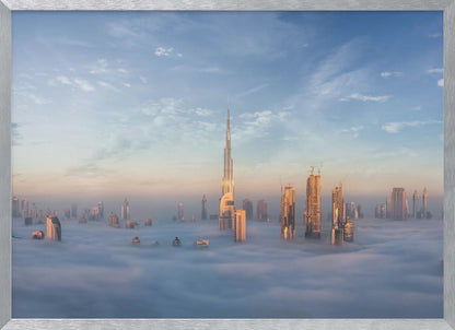 A framed photograph of the Dubai skyline, with the Burj Khalifa and other skyscrapers piercing through a thick blanket of low-lying clouds or fog. The scene is bathed in the warm, golden light of sunrise or sunset against a pale blue sky. Poster