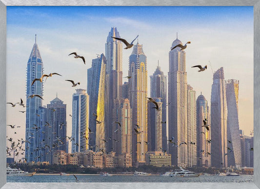 A framed artwork of the Dubai city skyline, featuring modern skyscrapers bathed in the golden light of sunset. A flock of seagulls flies in the foreground above the water, where several yachts are docked. Artwork