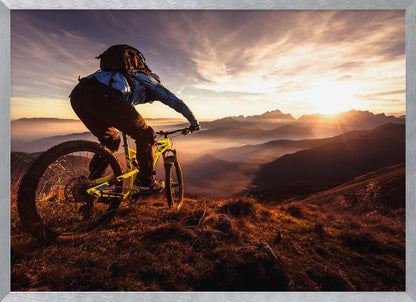A mountain biker, seen from behind, stops at the top of a grassy hill to admire a spectacular sunset over a vast mountain range. The valleys below are filled with mist, and the sun's golden rays create a stunning starburst effect against the silhouetted peaks. Print