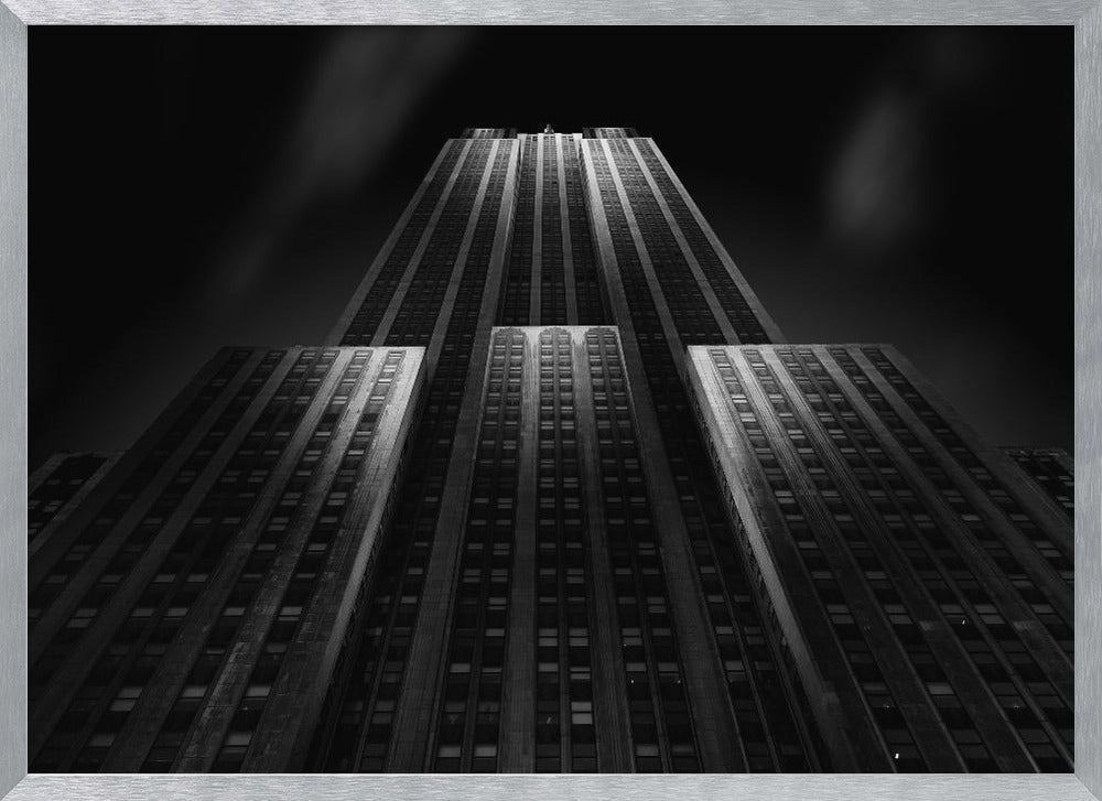 A dramatic, low-angle black and white photograph of the Empire State Building at night. The perspective looks up from the base, emphasizing the skyscraper's height and Art Deco details. Spotlights highlight the vertical lines of the building against a dark, moody sky, all enclosed in a silver frame. Poster