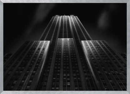 A dramatic, low-angle black and white photograph of the Empire State Building at night. The perspective looks up from the base, emphasizing the skyscraper's height and Art Deco details. Spotlights highlight the vertical lines of the building against a dark, moody sky, all enclosed in a silver frame. Poster