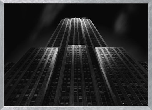 A dramatic, low-angle black and white photograph of the Empire State Building at night. The perspective looks up from the base, emphasizing the skyscraper's height and Art Deco details. Spotlights highlight the vertical lines of the building against a dark, moody sky, all enclosed in a silver frame. Poster