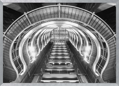 A symmetrical, low-angle black and white photograph of a futuristic escalator or grand staircase. The structure features illuminated, repeating curved arches on both sides, creating a tunnel-like perspective leading upwards. The image is presented within a silver-colored frame. Artwork