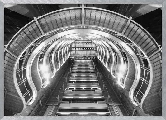 A symmetrical, low-angle black and white photograph of a futuristic escalator or grand staircase. The structure features illuminated, repeating curved arches on both sides, creating a tunnel-like perspective leading upwards. The image is presented within a silver-colored frame. Artwork