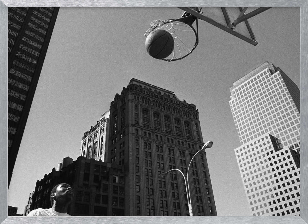 A low-angle, black and white photograph capturing a basketball as it's about to fall through the hoop, set against a backdrop of towering city skyscrapers. A player's face is visible in the bottom left corner, looking up at the ball. The entire image is presented within a silver-colored frame. Artwork