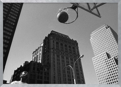 A low-angle, black and white photograph capturing a basketball as it's about to fall through the hoop, set against a backdrop of towering city skyscrapers. A player's face is visible in the bottom left corner, looking up at the ball. The entire image is presented within a silver-colored frame. Artwork