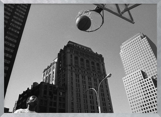 A low-angle, black and white photograph capturing a basketball as it's about to fall through the hoop, set against a backdrop of towering city skyscrapers. A player's face is visible in the bottom left corner, looking up at the ball. The entire image is presented within a silver-colored frame. Artwork