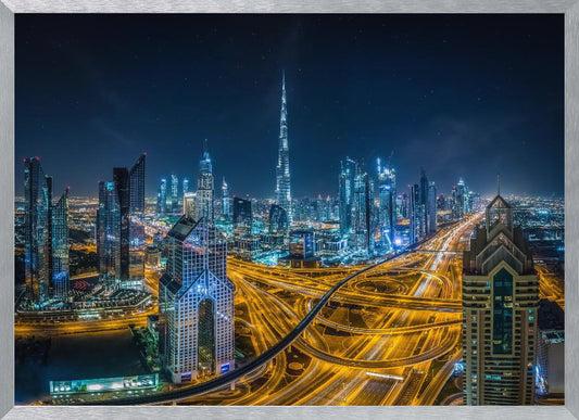 A vibrant aerial photograph of the Dubai skyline at night, with the iconic Burj Khalifa towering in the center. A complex web of brightly lit highways with light trails from traffic dominates the foreground, weaving between modern skyscrapers under a starry night sky. Decor
