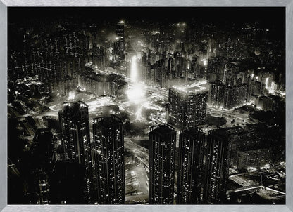 A framed black and white aerial photograph of a dense city at night. The high-angle view captures countless illuminated skyscrapers and winding highways, creating a dramatic and bustling urban landscape. Print