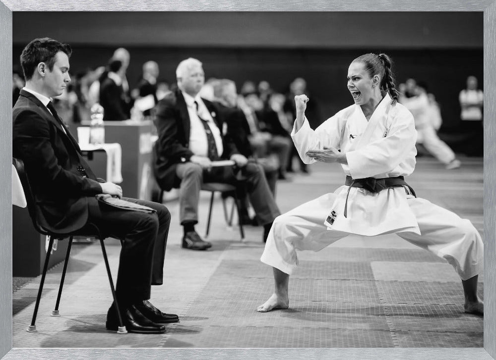 A dynamic black and white photograph of a female martial artist in a white gi and black belt, mid-kiai. She is in a deep, wide stance on a matted floor, her face filled with intensity and her fist clenched. In the foreground, a man in a suit sits and watches, while other spectators are seated in the blurred background, suggesting a competition or demonstration setting. The entire image has a silver-colored frame. Print