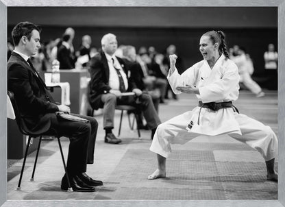 A dynamic black and white photograph of a female martial artist in a white gi and black belt, mid-kiai. She is in a deep, wide stance on a matted floor, her face filled with intensity and her fist clenched. In the foreground, a man in a suit sits and watches, while other spectators are seated in the blurred background, suggesting a competition or demonstration setting. The entire image has a silver-colored frame. Print