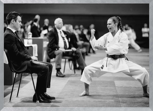 A dynamic black and white photograph of a female martial artist in a white gi and black belt, mid-kiai. She is in a deep, wide stance on a matted floor, her face filled with intensity and her fist clenched. In the foreground, a man in a suit sits and watches, while other spectators are seated in the blurred background, suggesting a competition or demonstration setting. The entire image has a silver-colored frame. Print