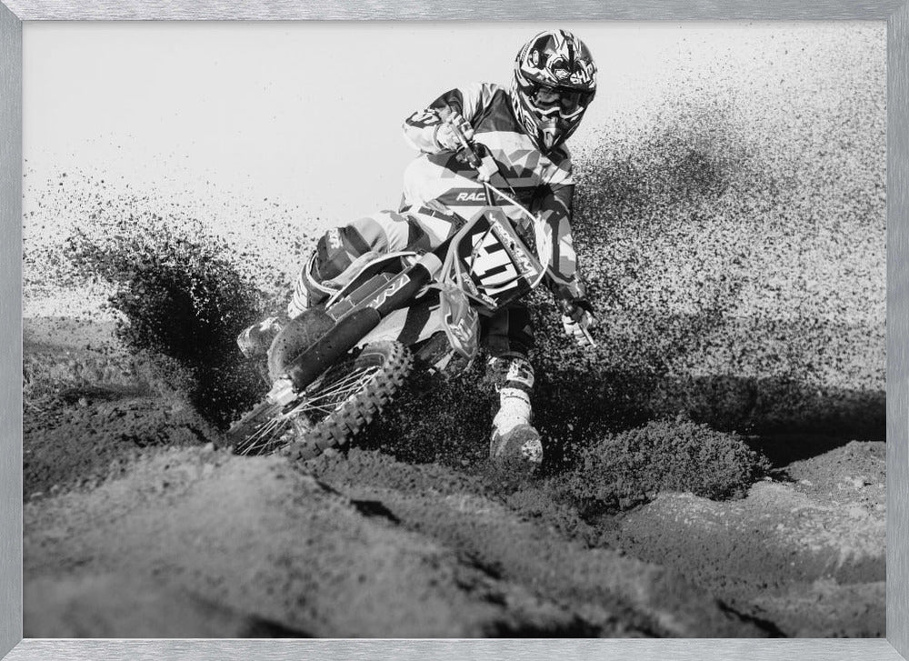 A dynamic black and white action photo of a motocross rider in full gear leaning hard into a turn, kicking up a large spray of dirt and sand from the track. The image is presented within a silver frame. Decor