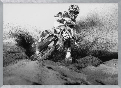 A dynamic black and white action photo of a motocross rider in full gear leaning hard into a turn, kicking up a large spray of dirt and sand from the track. The image is presented within a silver frame. Decor