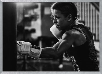 A dramatic black and white photo of a muscular female boxer in profile, intensely focused as she punches a heavy bag, set within a silver frame. Print