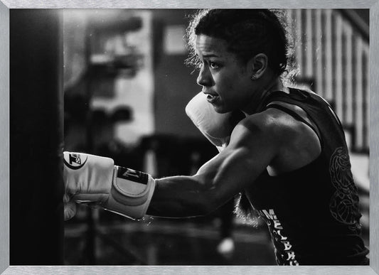 A dramatic black and white photo of a muscular female boxer in profile, intensely focused as she punches a heavy bag, set within a silver frame. Print