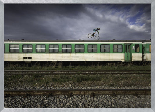 A BMX rider is captured in mid-air performing a stunt on the roof of a green and white passenger train car, set against a dramatic, stormy sky. The scene takes place on railway tracks with gravel and overgrown grass, and the entire image is enclosed in a silver frame. Decor