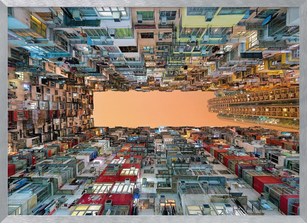An upward-looking photograph from a courtyard surrounded by densely packed, multi-colored apartment buildings that create a rectangular frame around an orange sky. The unique perspective creates a surreal and geometric view of an urban landscape, all enclosed in a silver frame. Artwork