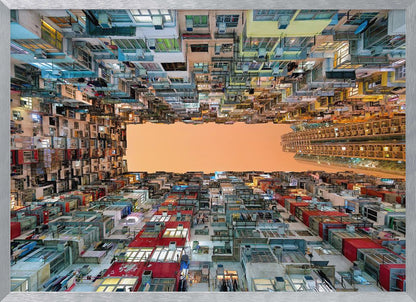 An upward-looking photograph from a courtyard surrounded by densely packed, multi-colored apartment buildings that create a rectangular frame around an orange sky. The unique perspective creates a surreal and geometric view of an urban landscape, all enclosed in a silver frame. Artwork