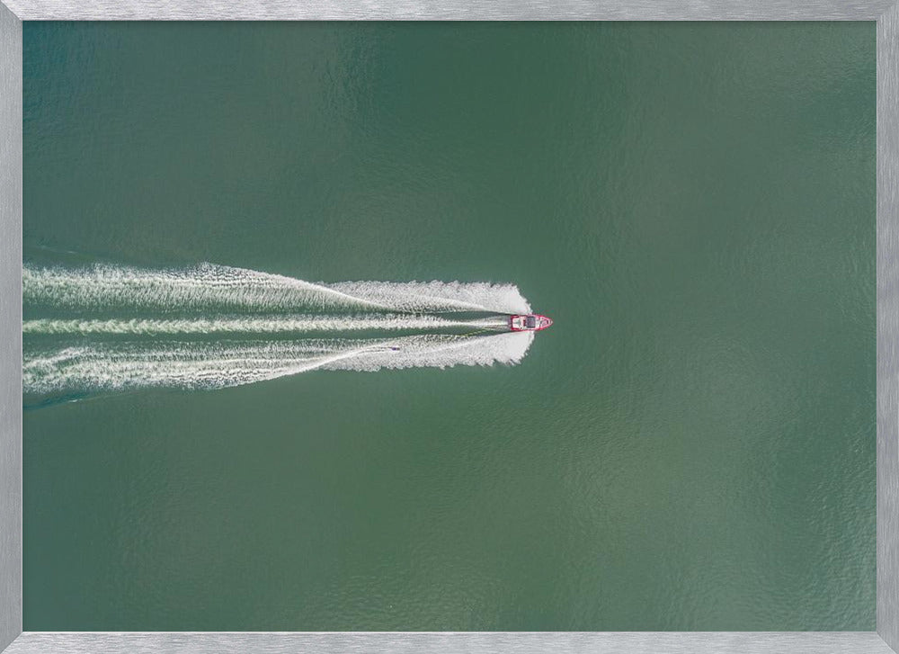 An aerial top-down photograph of a red motorboat speeding across a vast expanse of calm, green water, leaving a long, white, frothy wake behind it. The scene is presented within a simple silver frame. Print