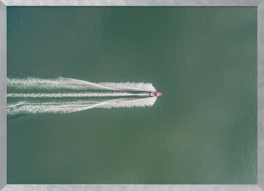 An aerial top-down photograph of a red motorboat speeding across a vast expanse of calm, green water, leaving a long, white, frothy wake behind it. The scene is presented within a simple silver frame. Print