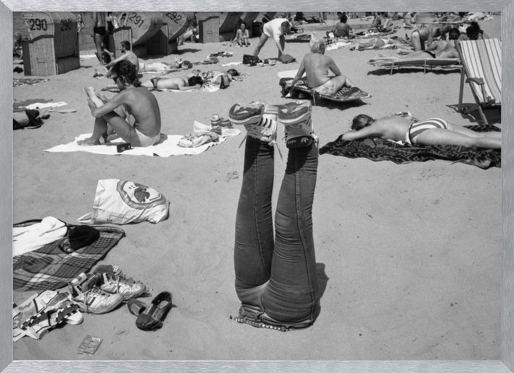 A quirky, vintage black and white photograph of a crowded beach. The central focus is a person lying on their back with their legs straight up in the air, oddly wearing jeans and sneakers. Surrounding them are numerous other beachgoers sunbathing on towels and in wicker beach chairs, creating a busy and candid summer scene. Wall Art