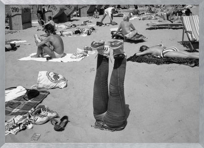 A quirky, vintage black and white photograph of a crowded beach. The central focus is a person lying on their back with their legs straight up in the air, oddly wearing jeans and sneakers. Surrounding them are numerous other beachgoers sunbathing on towels and in wicker beach chairs, creating a busy and candid summer scene. Wall Art