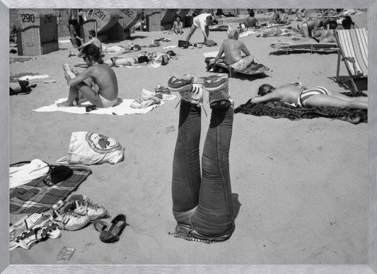 A quirky, vintage black and white photograph of a crowded beach. The central focus is a person lying on their back with their legs straight up in the air, oddly wearing jeans and sneakers. Surrounding them are numerous other beachgoers sunbathing on towels and in wicker beach chairs, creating a busy and candid summer scene. Wall Art