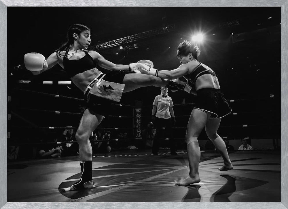 A dramatic black and white action photograph of two female kickboxers in a ring. One fighter, airborne, executes a high kick that connects with the other fighter's gloves, who is bracing for the impact. A referee stands in the background under bright arena lights. Poster