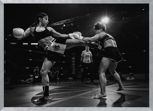 A dramatic black and white action photograph of two female kickboxers in a ring. One fighter, airborne, executes a high kick that connects with the other fighter's gloves, who is bracing for the impact. A referee stands in the background under bright arena lights. Poster