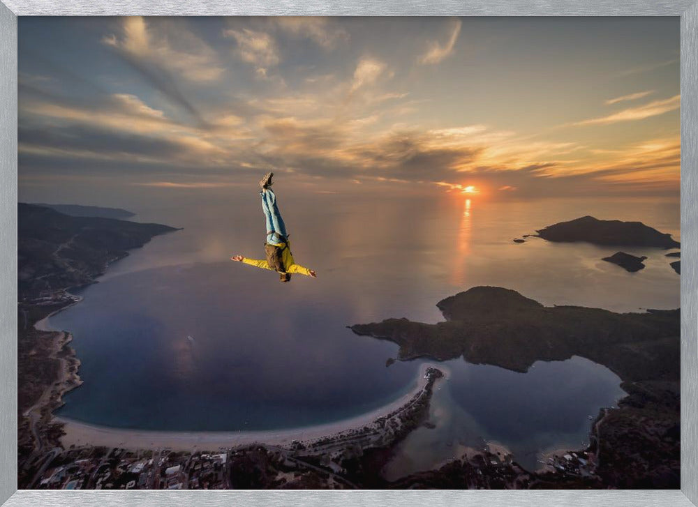 A breathtaking aerial view of a skydiver in a head-down position, freefalling over a vast coastal bay at sunset. The setting sun casts a golden glow over the ocean and clouds, highlighting the islands and coastline below. Decor