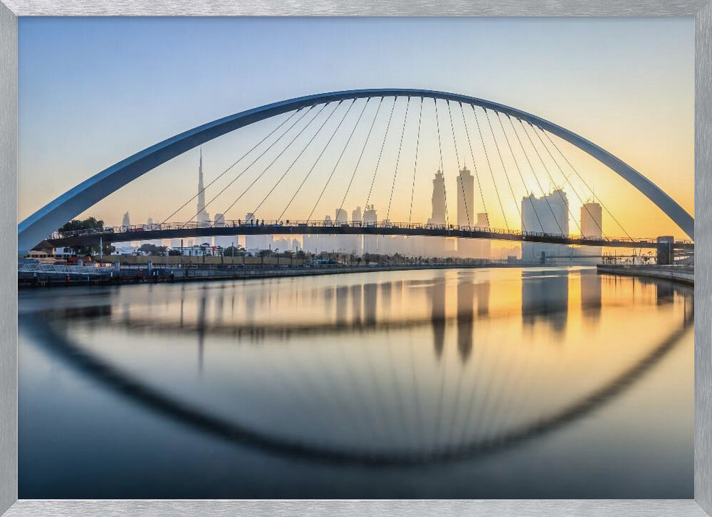 A framed photograph of the Dubai Water Canal Bridge at sunrise, with its large arch spanning across the water. The Dubai skyline, including the Burj Khalifa, is visible in the background, silhouetted against a golden and blue sky. The bridge and buildings create a perfect reflection on the calm surface of the water. Wall Art