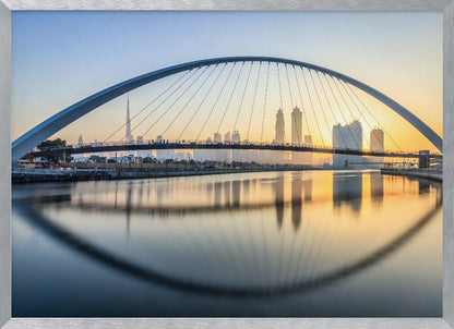 A framed photograph of the Dubai Water Canal Bridge at sunrise, with its large arch spanning across the water. The Dubai skyline, including the Burj Khalifa, is visible in the background, silhouetted against a golden and blue sky. The bridge and buildings create a perfect reflection on the calm surface of the water. Wall Art
