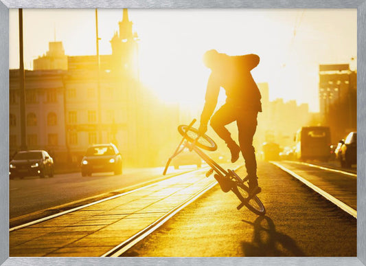 A silhouetted BMX rider performs a trick on a city street during a brilliant golden sunset, with the sun flaring behind them and casting long shadows on the tram tracks. Decor