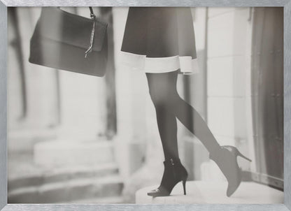 A framed, black and white fashion photograph focusing on the lower half of a woman as she walks. She is wearing a dark skirt with a white hem, black tights, and stylish high-heeled ankle boots, and she is carrying a dark handbag. Print