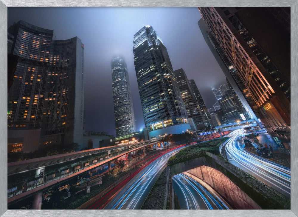 A dynamic, low-angle, long-exposure shot of a modern city at night. Towering skyscrapers with illuminated windows pierce through a foggy, dark blue sky. Below, curving highways are filled with the vibrant red and white light trails of moving traffic. Print