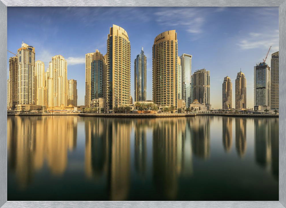 A panoramic photograph of the Dubai Marina skyline with its golden skyscrapers reflecting in the calm, dark water below, all under a clear blue sky and framed in silver. Wall Art